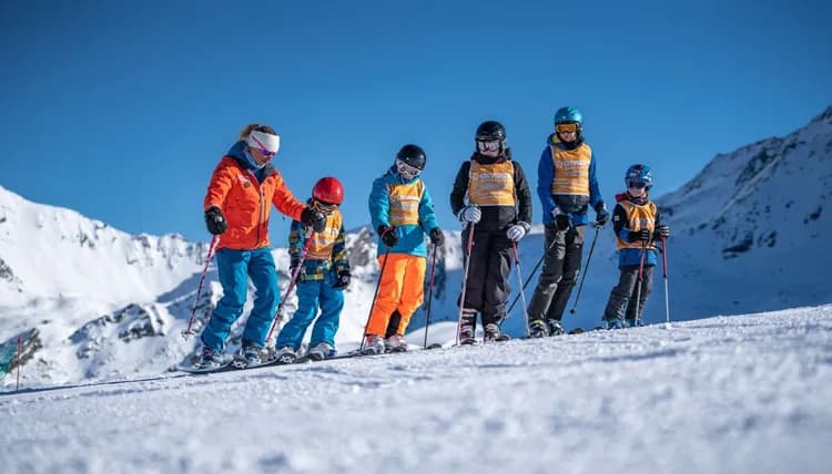 Grupo de alumnos con esquís, cascos y petos de escuela de esquí, alineados en una pista nevada junto a su monitora, con montañas y cielo azul al fondo