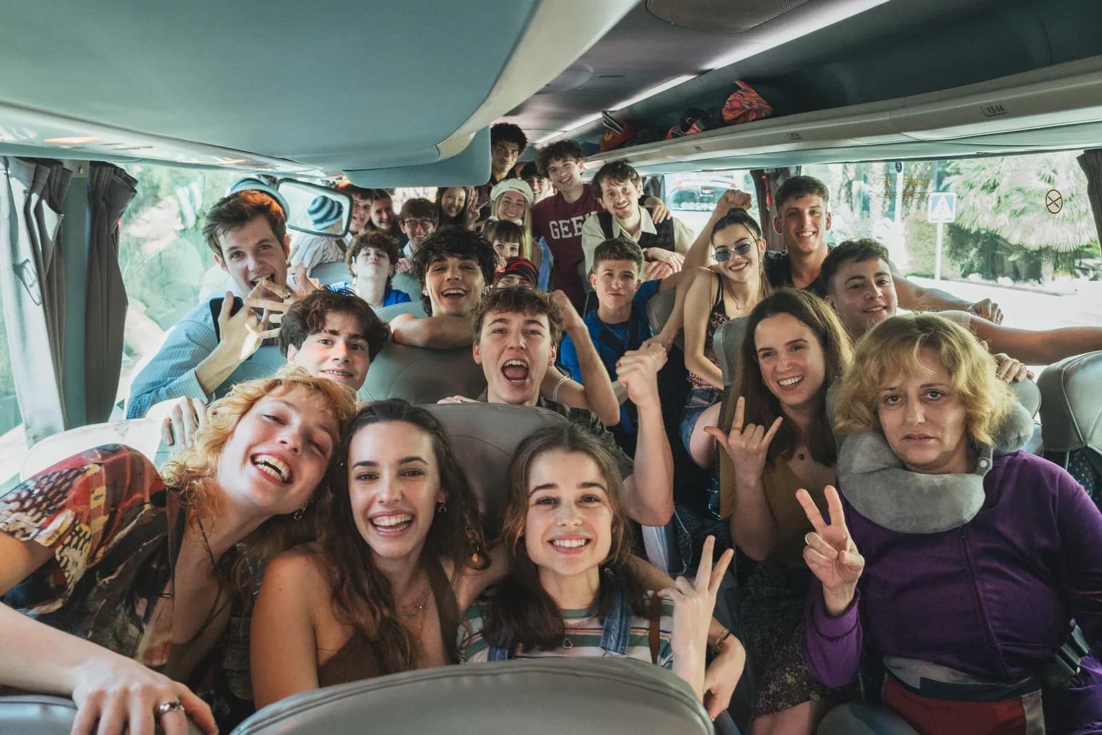 Grupo grande de estudiantes de viaje de fin de curso apiñados en los asientos del autocar, riendo y haciendo el signo de la victoria hacia la cámara